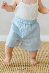 Toddler standing in Avauma Sunny Stripe Pants Blueberry Powder. Child wearing light blue striped shorts and a white top, ready for play.